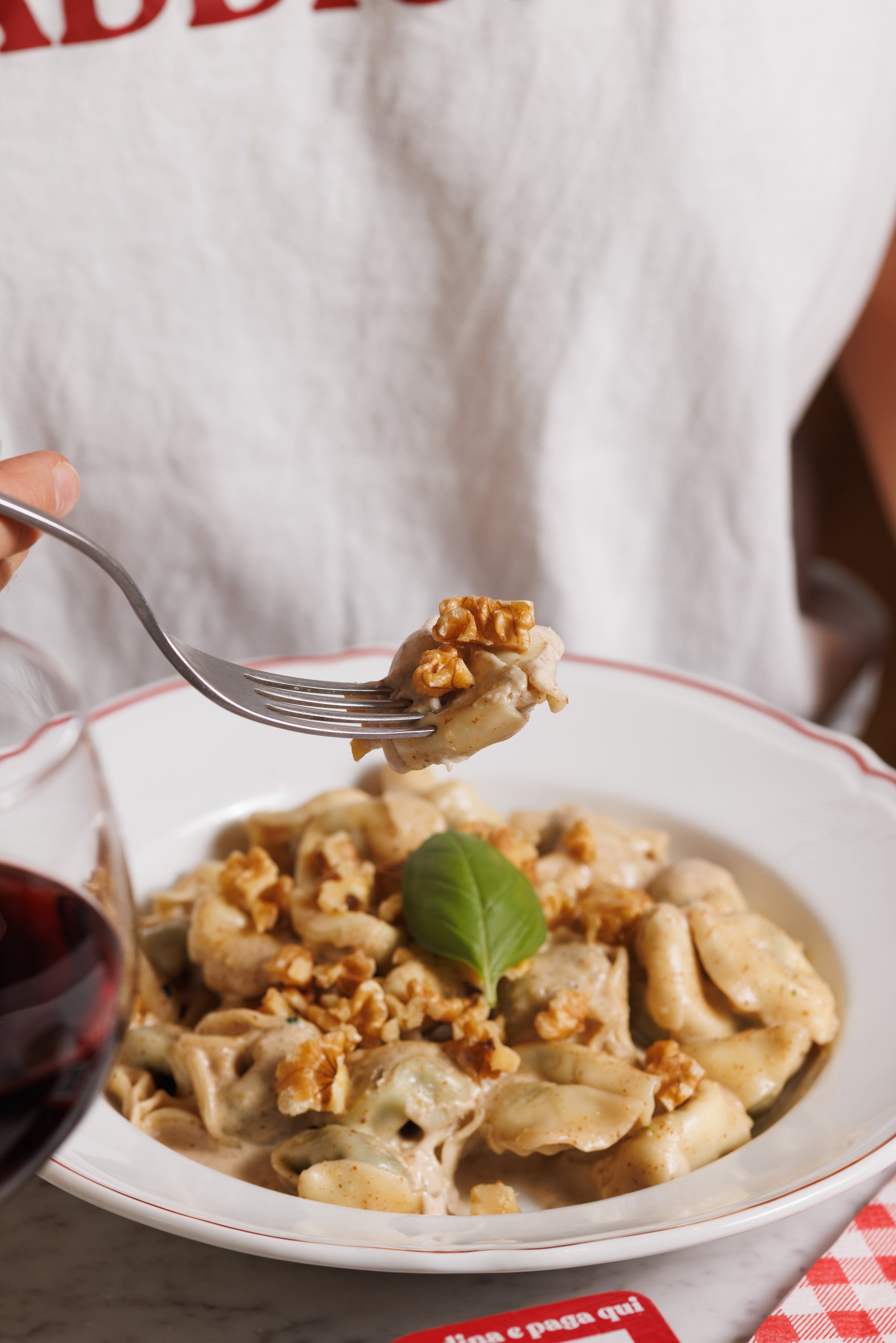 A close-up of a fork lifting a piece of creamy walnut tortellini from a plate, accompanied by a glass of red wine.
