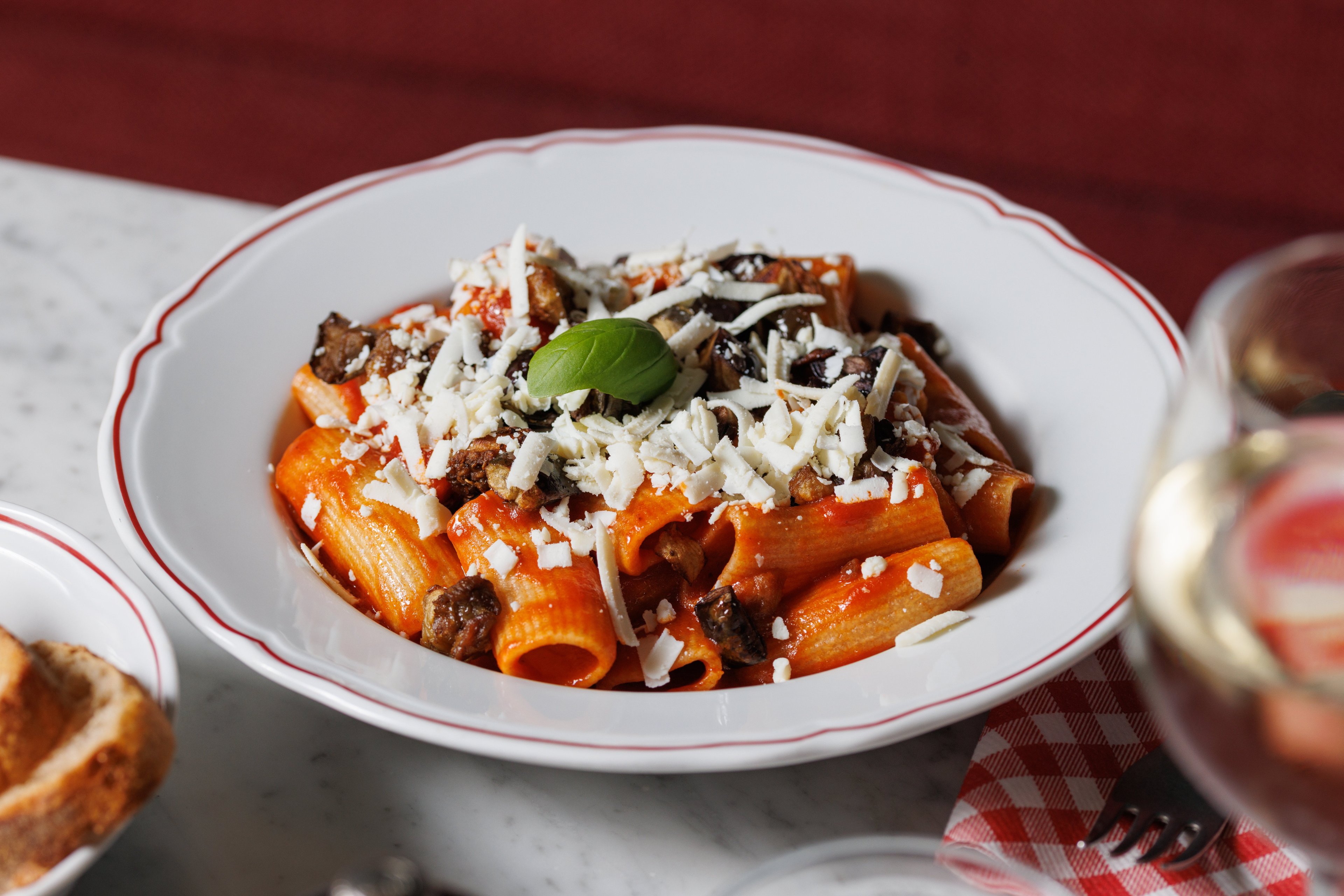 A plate of rigatoni pasta with tomato sauce, eggplant, and grated cheese is served on a marble table in a warm, inviting restaurant setting.