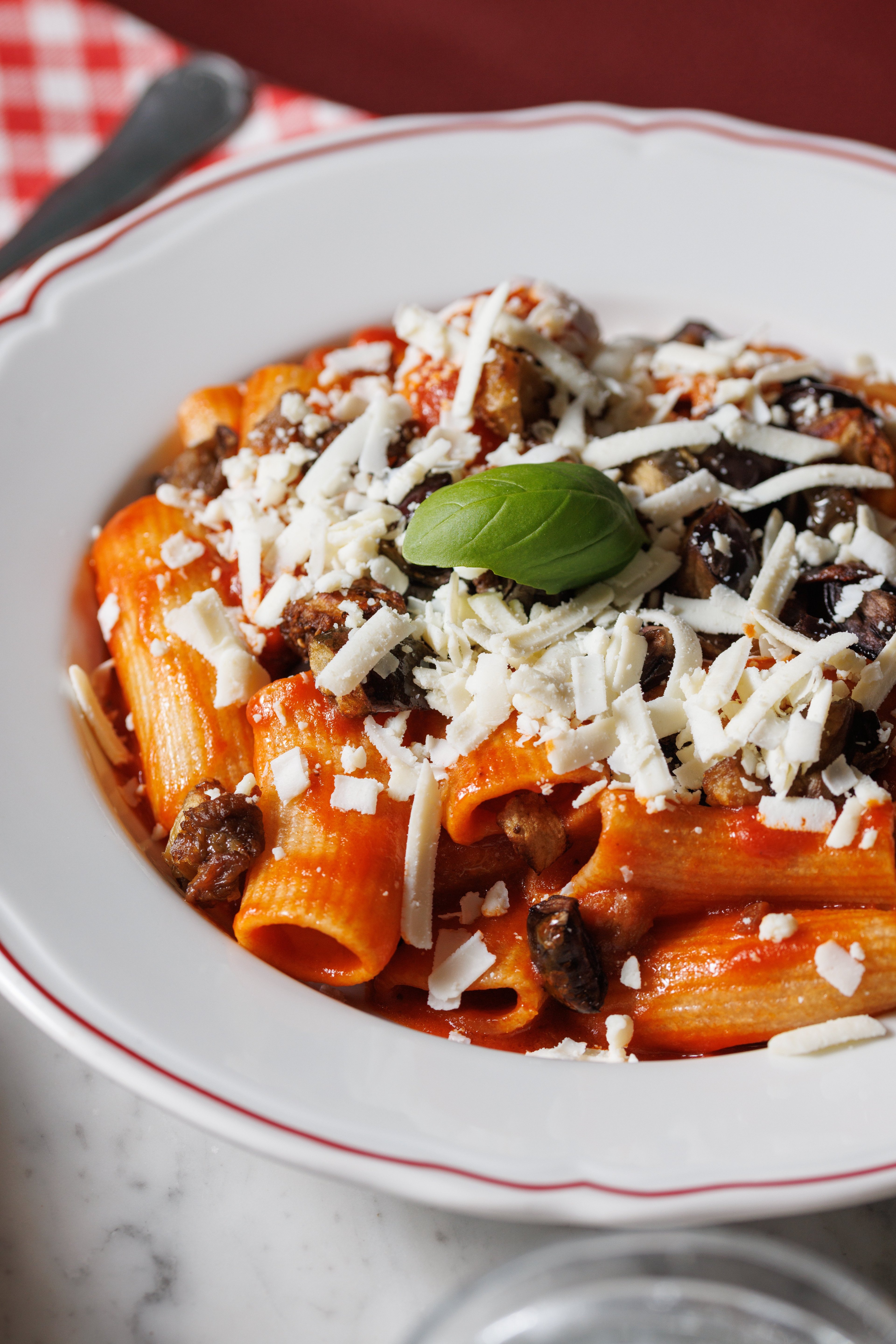 A close-up shot of a bowl of rigatoni pasta topped with tomato sauce, roasted eggplant, and shredded cheese.