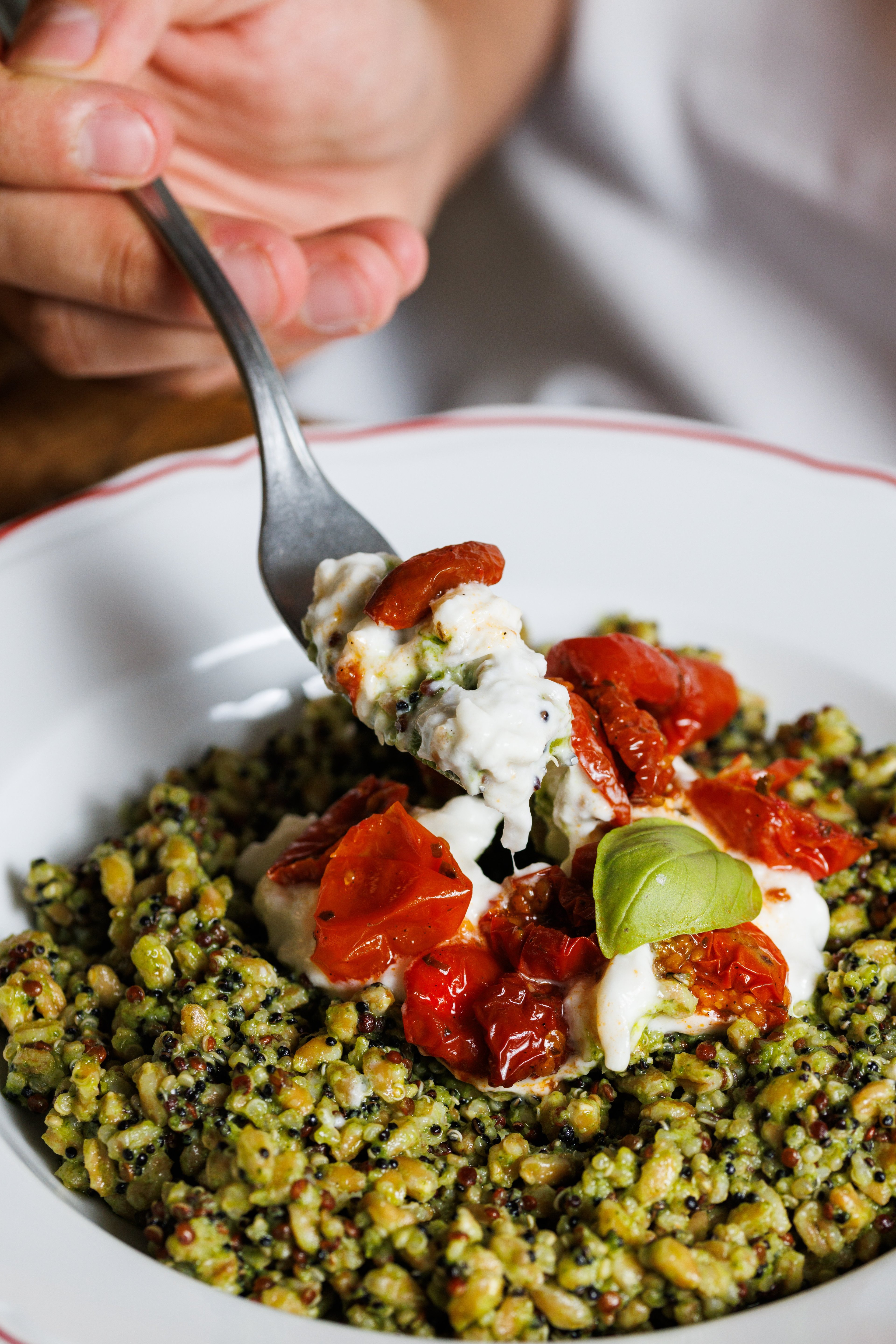 A close-up of a fork lifting a bite of creamy burrata cheese topped with sun-dried tomatoes over a bed of pesto-infused grain salad.