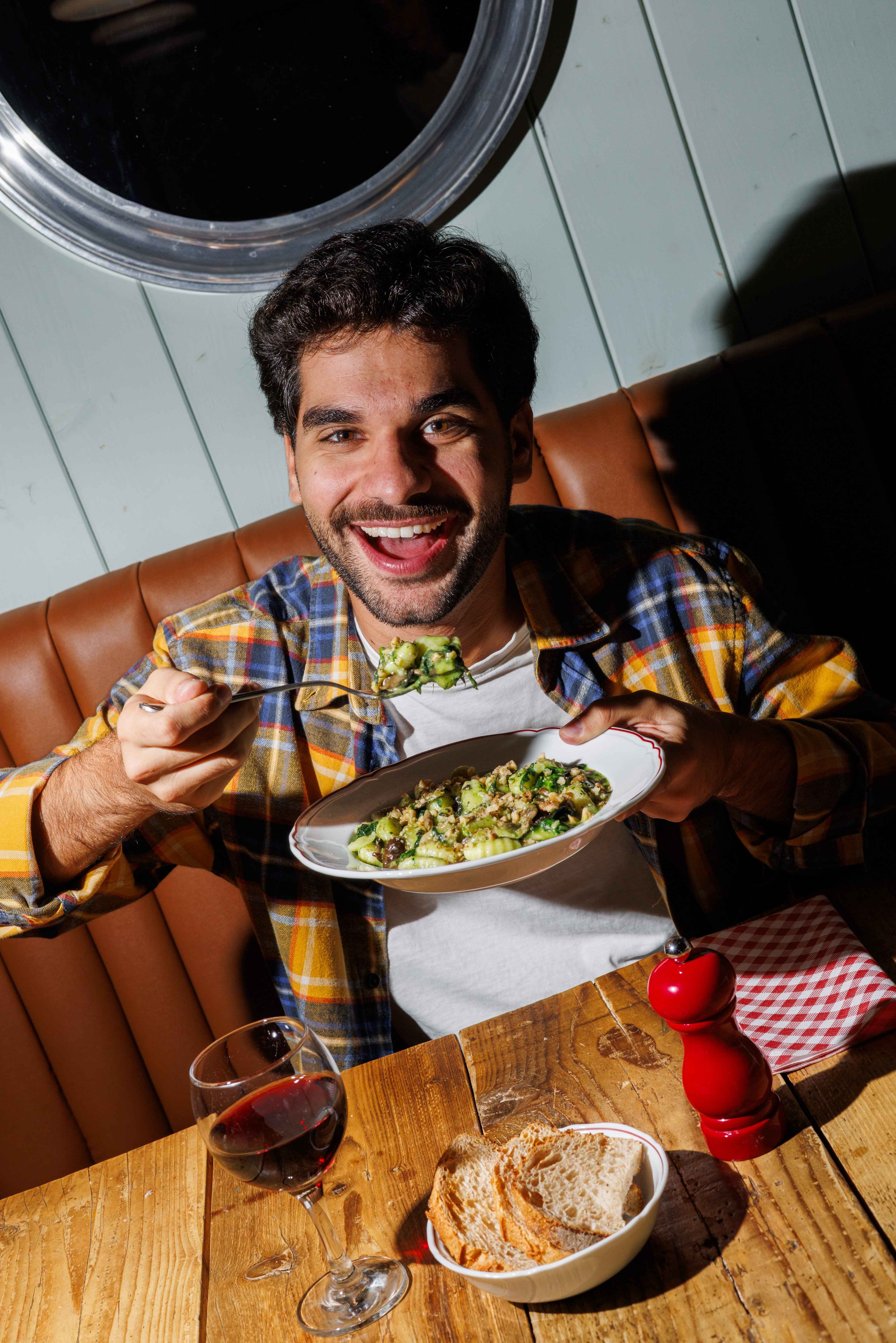 A cheerful man enjoys a plate of gnocchi with pesto and walnuts in a cozy, warmly lit restaurant.