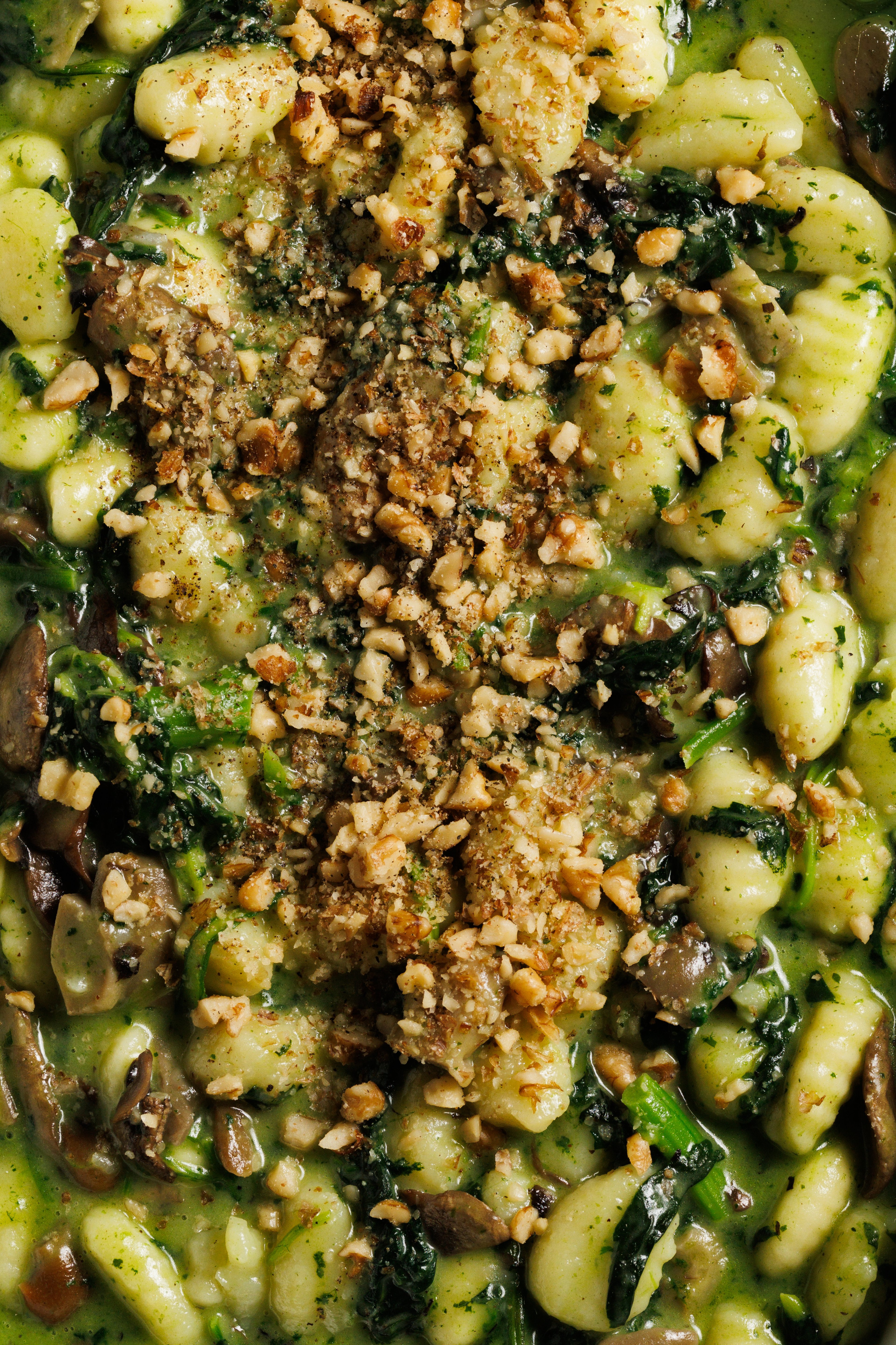 A close-up overhead shot of a creamy gnocchi dish topped with chopped walnuts, sautéed mushrooms, and fresh spinach.